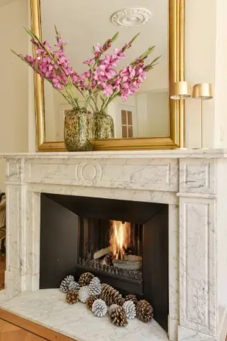 A marble fireplace with a lit fire, featuring a stylish fireplace surround decorated with pinecones on the hearth and two vases of pink flowers on the mantel, topped by a large gold-framed mirror above.