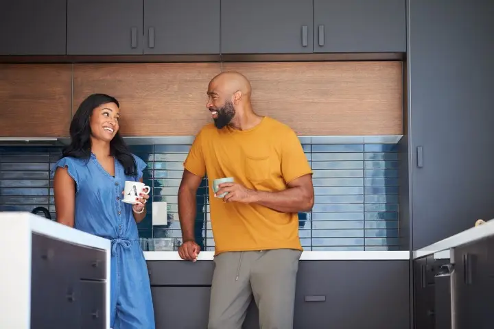 Two people stand in a modern kitchen, smiling at each other while having a conversation, surrounded by sleek cabinets and elegant stone countertops.