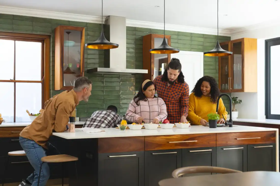 Four people preparing food together in a modern kitchen with a green tiled wall and dark cabinets. Two pendant lights hang above the countertop.