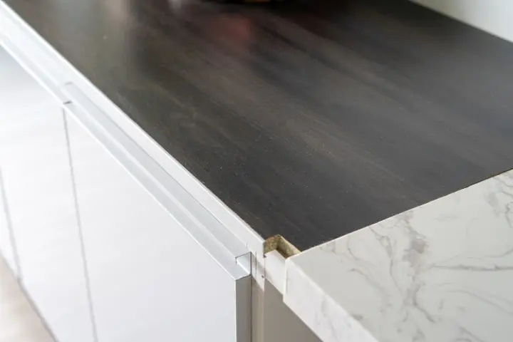 A kitchen countertop with a damaged edge profile exposes particle board beneath the dark laminate surface, situated beside a white marble-patterned countertop.