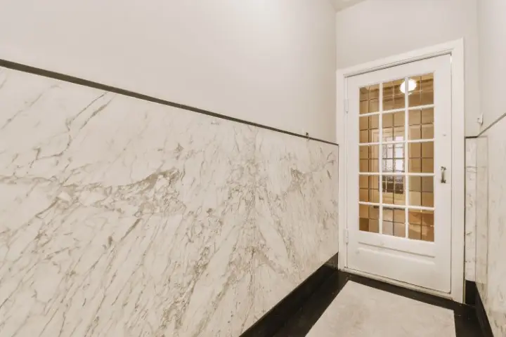 A hallway featuring modern luxury with white marble-patterned stone accent walls leads to a white-framed glass door, through which a light fixture is visible.