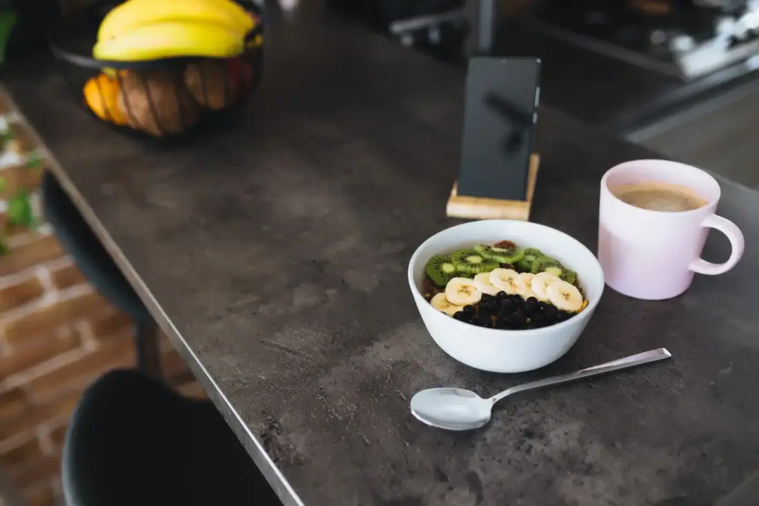 A bowl of cereal with sliced bananas and kiwi rests on a matte countertop, next to a glossy pink mug and smartphone. A spoon lies nearby, while in the background, a fruit basket adds charm to the kitchen’s stylish debate: matte vs. polished countertops.