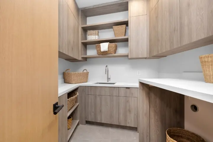 Modern laundry room with functional design features, including light wood laundry room cabinets, white countertops, open shelves, wicker baskets, and a sink under a wall-mounted faucet that elevates your space.