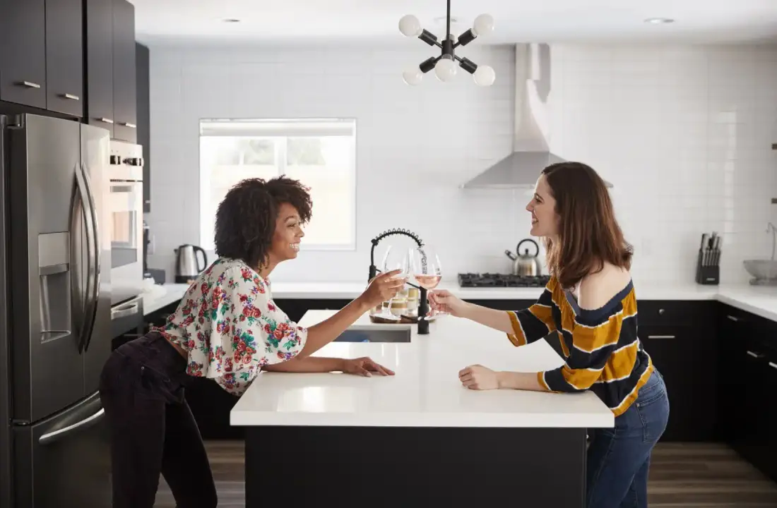 Two women stand in a modern kitchen, smiling and clinking wine glasses over a sleek island countertop, discussing the pros and cons of matte vs. polished finishes.