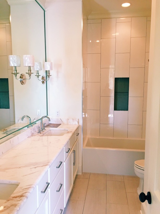 A bathroom features a large mirror above a marble countertop and twin sinks, accented by a stylish light fixture. The shower area, complete with tiled walls and a bathtub, harmoniously complements the sleek granite template design throughout.