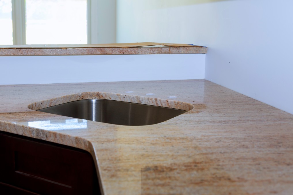 Marble countertop installation transforms any space, but here we feature a stunning granite kitchen countertop with an inset stainless steel sink, light-colored with brown veining, perfectly situated beneath a window.