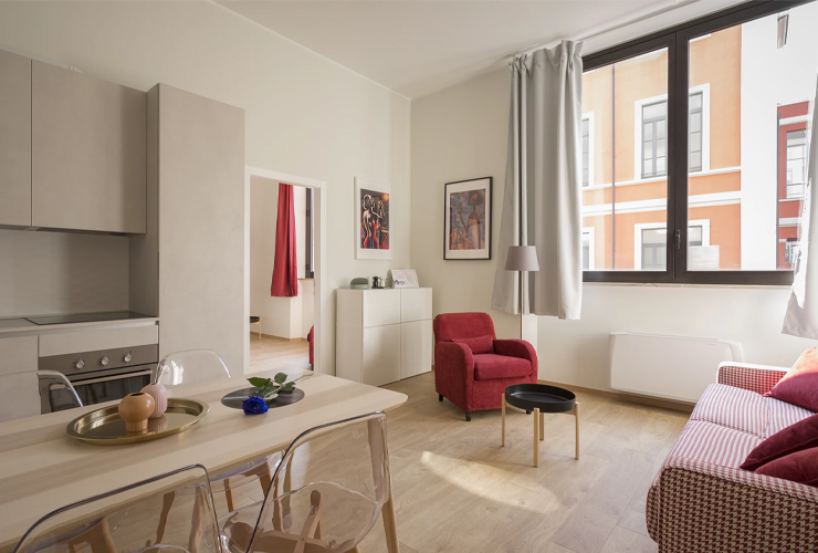 A modern living room with a red chair, beige sofa, and small black table. A kitchen area and dining table with clear chairs are visible. Large windows allow natural light in.