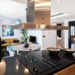 Modern kitchen with a black countertop and stainless steel range hood, overlooking a living room with a beige sofa and a round dining table with a flower vase.