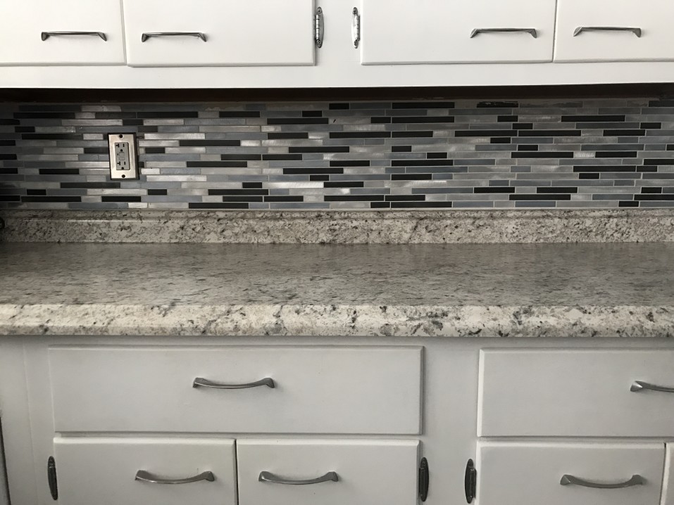 A kitchen countertop with a speckled gray marble design, complemented by white cabinets and a backsplash featuring rectangular gray and black tiles. An electrical outlet is visible on the wall.