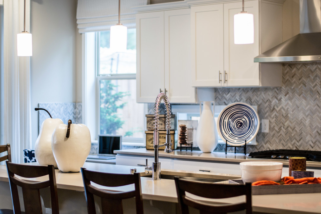Modern kitchen with white cabinets, a central island featuring marble countertop installation, decorative pear sculptures, a spiral-patterned plate, pendant lights, and a window view of foliage.