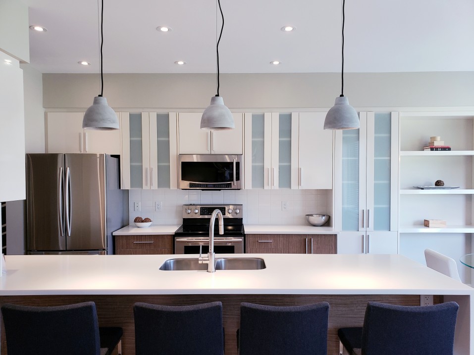 Modern kitchen with a large island featuring a sleek quartz template, pendant lights, stainless steel appliances, and white cabinets. Four dark chairs are arranged along the island countertop.