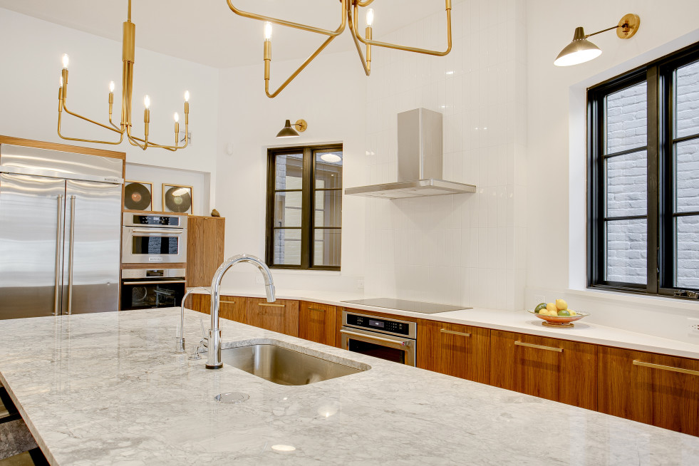 Modern kitchen with a seamlessly installed marble island, stainless steel appliances, wooden cabinets, and brass light fixtures. A bowl of fruit sits on the counter near the window.