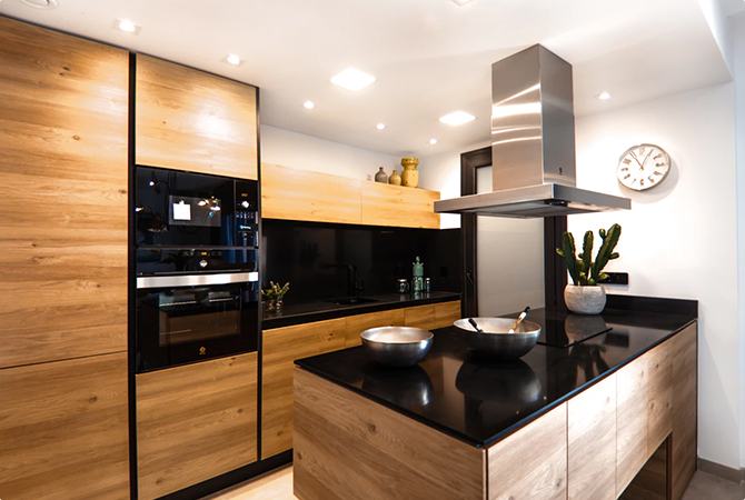 Modern kitchen with wooden cabinetry, black countertops, an island with two bowls, a built-in oven, and a range hood. A wall clock and plants add accents to the space.