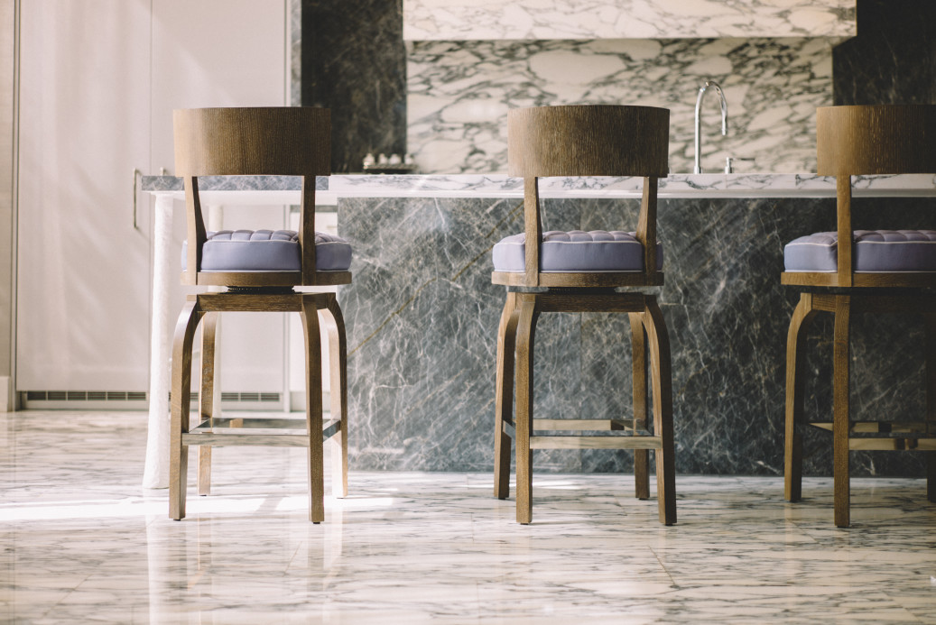 Three wooden bar stools with cushioned seats are positioned at a marble kitchen counter, showcasing expert marble countertop installation in a well-lit space with matching marble flooring.