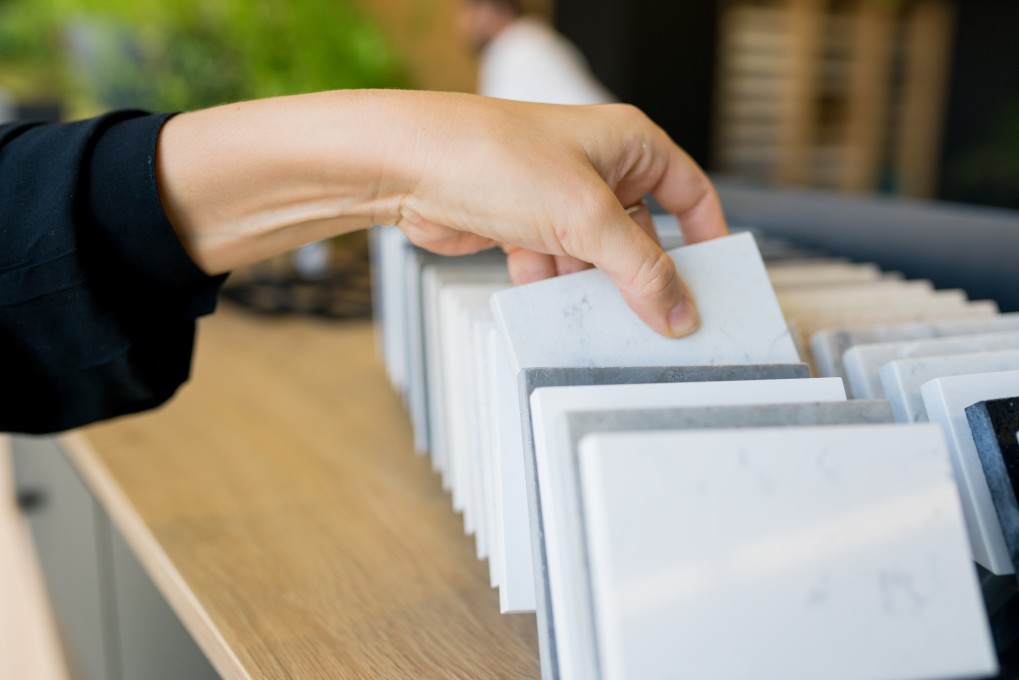 A person chooses a sample from a range of tile samples on a wooden surface, considering the quartz template for inspiration.