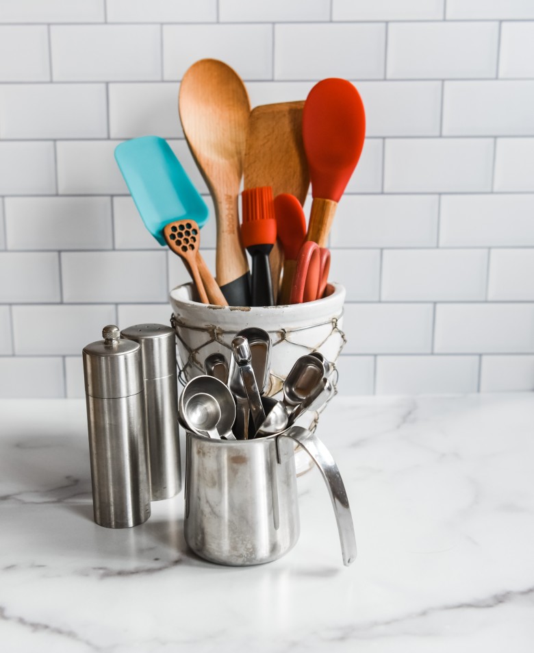 A kitchen countertop with a marble surface serves as the perfect template for culinary creativity, showcasing a container of wooden and silicone utensils alongside metal measuring spoons in a small jug, all set against a stylish tiled backsplash.