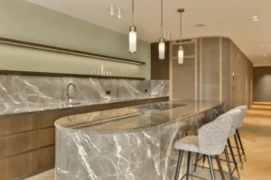 Modern kitchen with a marble island and premium countertops, bar stools, a sink, and hanging lights. Wooden cabinets and shelves line the walls.