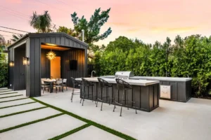 An outdoor kitchen featuring the best outdoor countertops for Florida, complete with a grill, bar seating, and dining area under a modern pergola. Surrounded by greenery, the space boasts a concrete floor with grass inlays, set against a stunning sunset sky.