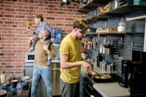 Two men and a child are in a kitchen with brick walls. One man holds the child on his shoulders, while the other cooks at the stove. Various kitchen items are visible on shelves.