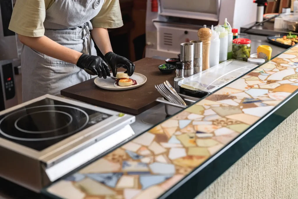 A person wearing black gloves prepares a dish on a kitchen countertop, surrounded by various cooking items and ingredients.