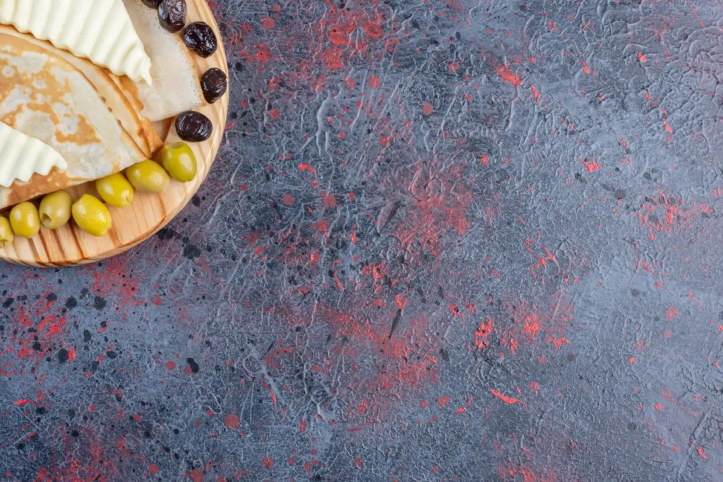 A wooden plate with cheese, meats, olives, and a flatbread rests on a textured blue and red background, reminiscent of a vibrant kitchen countertop color.