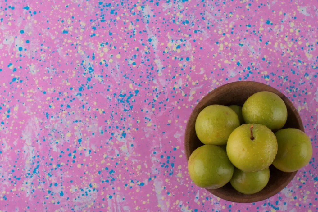 A wooden bowl brimming with green plums rests elegantly on a speckled pink countertop, adding a vibrant touch to the kitchen decor.