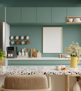 A modern kitchen features teal cabinets and a coffee maker, complemented by a terrazzo-patterned countertop. A stylish mug rack hangs nearby, while a blank picture frame and a vase of flowers add charm to the kitchen counter's delightful scene.