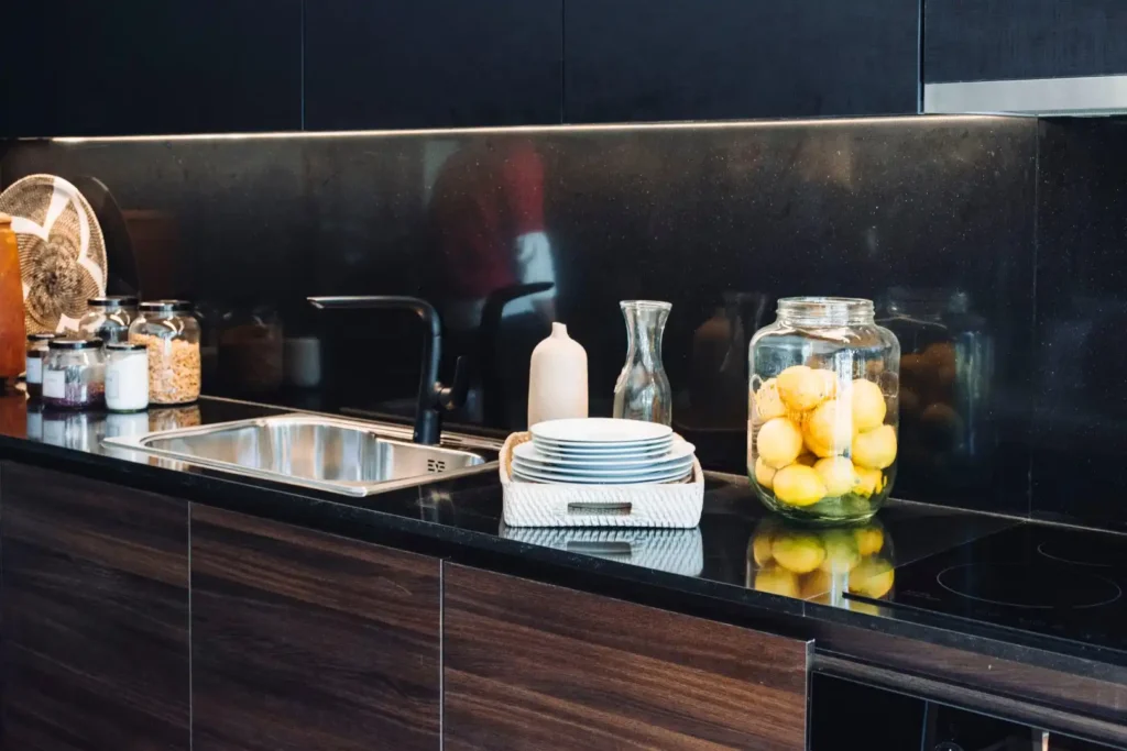 Modern kitchen countertop with a sink, black backsplash, and stacked plates showcases the perfect blend of matte vs. polished textures, complemented by a glass jar of lemons and various jars.