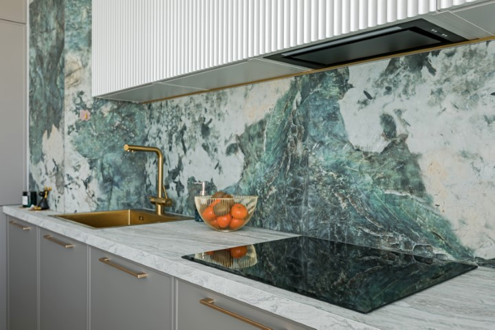 Modern kitchen with stone countertops and a marbled green and white backsplash, gold faucet, stovetop, and a basket of oranges on the countertop for a cohesive look.