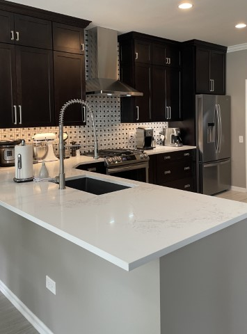 A modern kitchen featuring dark cabinets, stainless steel appliances, and white stone countertops. The mosaic tile backsplash adds a touch of elegance, enhancing the cohesive look. Complete with a sleek faucet, oven, refrigerator, and an array of cutting-edge kitchen gadgets.