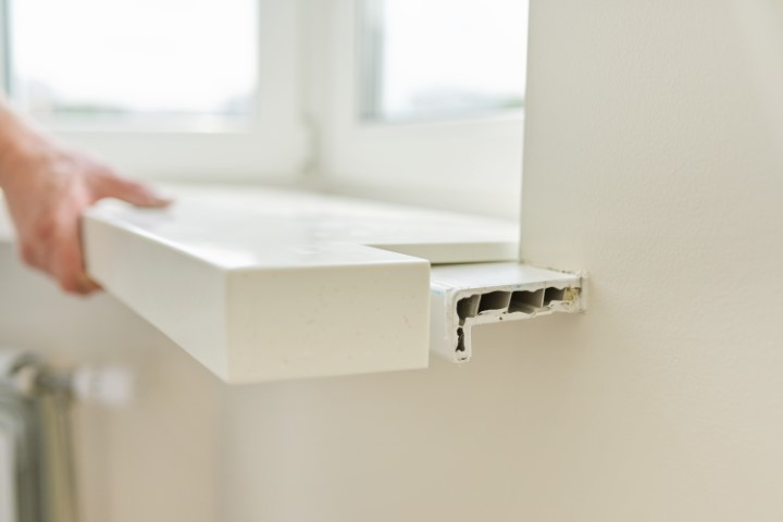 A person removes a white plastic window sill from its position beneath a window, exposing its hollow interior and the wall beneath—similar to examining the hidden structure of kitchen countertops or a countertop edge profile.