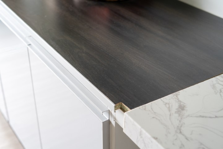 A kitchen countertop with a damaged edge profile exposes particle board beneath the dark laminate surface, situated beside a white marble-patterned countertop.