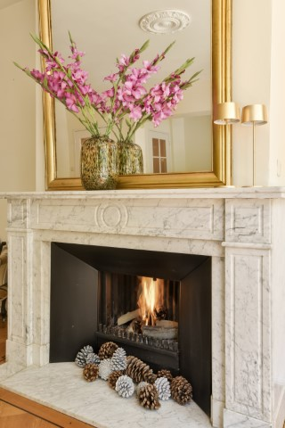 A marble fireplace with a lit fire, featuring a stylish fireplace surround decorated with pinecones on the hearth and two vases of pink flowers on the mantel, topped by a large gold-framed mirror above.