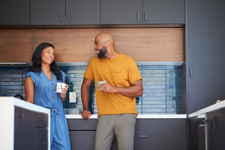 Two people stand in a modern kitchen, smiling at each other while having a conversation, surrounded by sleek cabinets and elegant stone countertops.