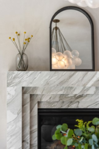A stylish fireplace with a marble surround features a glass vase of yellow flowers, a black-framed mirror, and a modern chandelier reflected in the mirror.