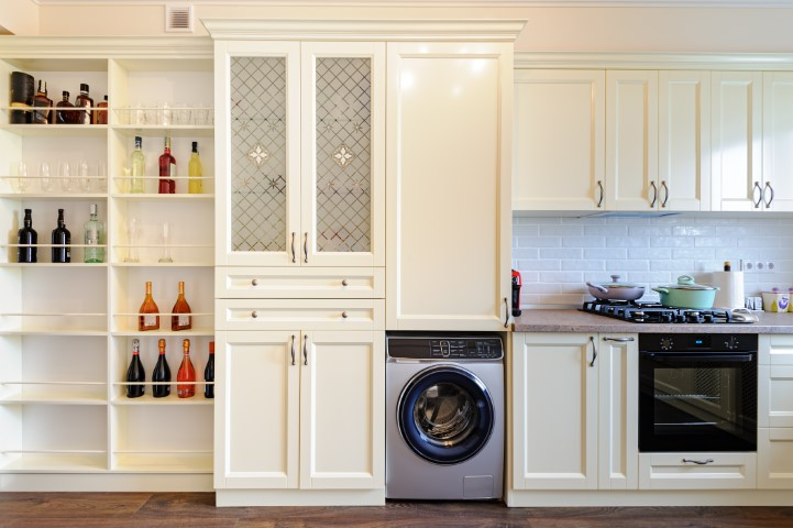 A modern kitchen with cream cabinets and open shelves holding bottles features a built-in washing machine, laundry room cabinets, a gas stove, and an oven under a tiled backsplash for functional design.