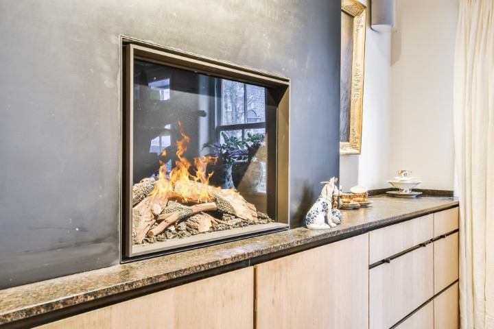 Modern glass-enclosed fireplace with a visible fire, showcasing a sleek Fireplace Style set in a black wall above a stone countertop with cabinets and decorative items nearby.