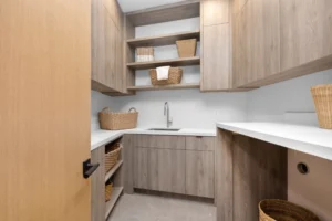 Modern laundry room with functional design features, including light wood laundry room cabinets, white countertops, open shelves, wicker baskets, and a sink under a wall-mounted faucet that elevates your space.